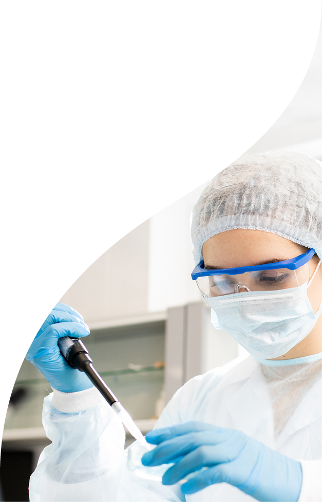 Serious concentrated female microbiologist in sterile clothing and safety goggles sitting at table and dropping reagent in petri dish while doing research in laboratory